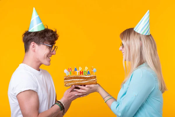 Zabawna Młoda para w czapki papierowe i ciasto zrobić głupie twarz i życzymy szczęśliwych urodzin stojąc na żółtym tle. Koncepcja gratulacji i oszukiwanie wokół. — Zdjęcie stockowe