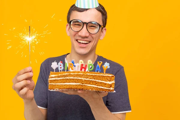 Retrato de um cara positivo engraçado com um boné de papel e óculos segurando um bolo caseiro congratulatório em suas mãos em um fundo amarelo. Conceito e diversão e celebração . — Fotografia de Stock