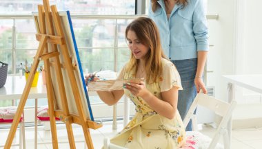 Sanat sınıfı, çizim ve yaratıcılık kavramı - Palet ve boya fırçası ile şövale önünde oturan kız öğrenciyakın çekim. Öğretmen ona yardım ediyor.