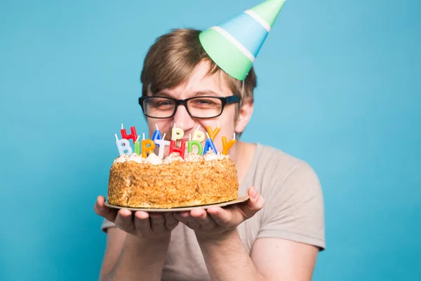Szalony zabawny młody człowiek w okularach i papier gratulacyjny kapelusze gospodarstwa ciasta szczęśliwy urodziny stojący na niebieskim tle. — Zdjęcie stockowe