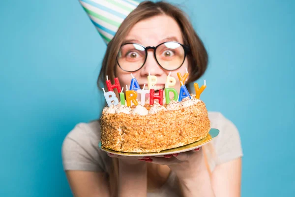 Charming merry crazy young girl student in congratulatory paper hat holding a happy birthday cake in her hands standing against a blue background. Advertising space. Royalty Free Stock Images