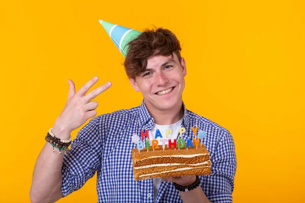 Engraçado cara positivo segura em suas mãos um bolo caseiro com a inscrição feliz aniversário posando em um fundo amarelo. Conceito de feriados e aniversários . — Fotografia de Stock