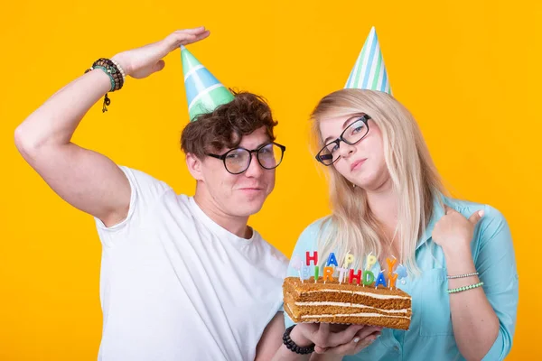 Pareja joven divertida en gorras de papel y con un pastel hacen una cara tonta y desean feliz cumpleaños mientras están de pie sobre un fondo amarillo. Concepto de felicitaciones y bromas . —  Fotos de Stock