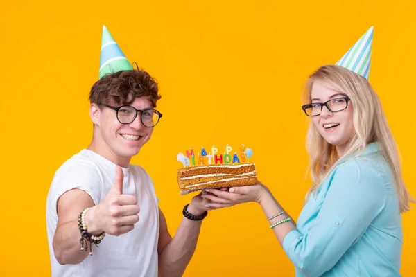 Komik nerd adam ve kadın tatil kapaklar ve gözlük sarı arka plan üzerinde mumlar ile doğum günü pastası tutan giyiyor — Stok fotoğraf