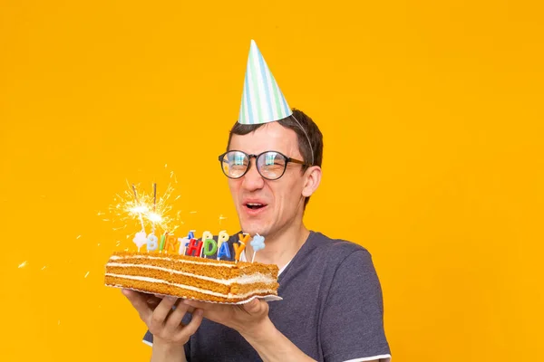 キャップと燃えるキャンドルと黄色の背景にポーズをとる彼の手の中に自家製ケーキを持つ肯定的な面白い若いアジアの男。記念日と誕生日のコンセプト. — ストック写真
