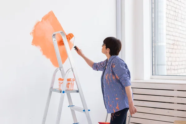 漂亮的微笑中年妇女用油漆滚筒在家里的内墙上画画。重新装修、翻新、公寓维修和茶点概念. — 图库照片