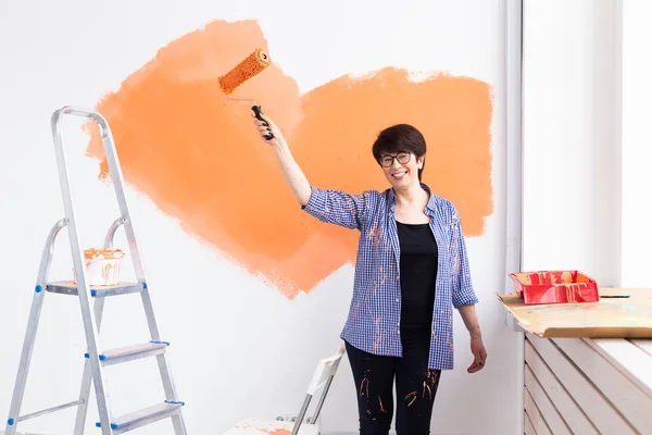 Feliz mujer de mediana edad pintando la pared interior con rodillo de pintura en casa nueva. Una mujer con rodillo aplicando pintura en una pared. — Foto de Stock