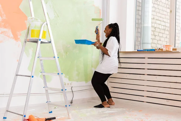 Felice donna afro-americana sorridente pittura parete interna della nuova casa. Ristrutturazione, ristrutturazione, riparazione di appartamenti e concetto di ristoro . — Foto Stock