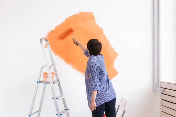 Feliz mujer de mediana edad pintando la pared interior con rodillo de pintura en casa nueva. Una mujer con rodillo aplicando pintura en una pared. —  Fotos de Stock