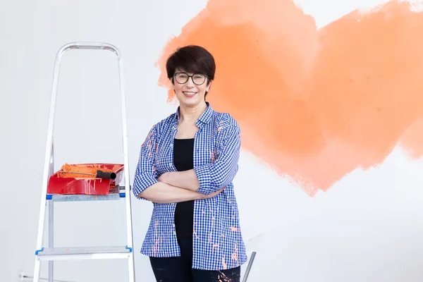 Mooie glimlachende vrouw van middelbare leeftijd die interieur muur van huis schildert met verfroller. Herinrichting, renovatie, appartement reparatie en verfrissing concept. — Stockfoto