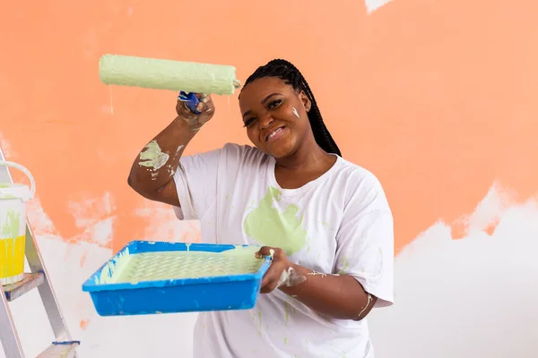 Güzel gülümseyen Afro-Amerikan kadın evin içini boya rulosuyla boyuyor. Dekorasyon, yenileme, apartman onarımı ve ferahlatıcı konsept. — Stok fotoğraf