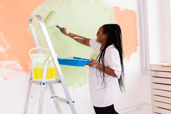 Destul de zâmbitor african american femeie pictura perete interior de casă cu role de vopsea. Redecorare, renovare, reparații apartamente și concept de băuturi răcoritoare . — Fotografie, imagine de stoc