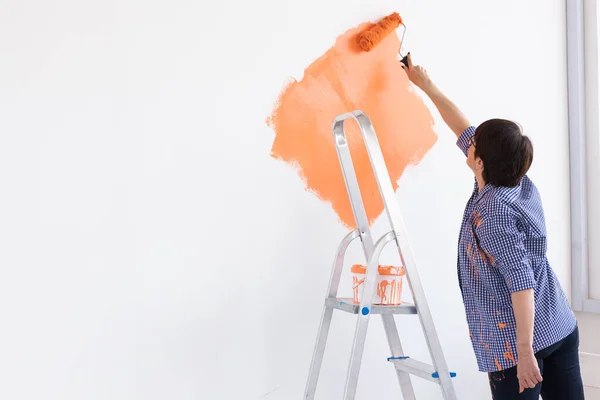 Yeni evde orta yaşlı mutlu bir kadın boya rulosuyla duvarları boyuyor. Duvara boya süren tekerlekli sandalyesi olan bir kadın. Boşluğu kopyala. — Stok fotoğraf