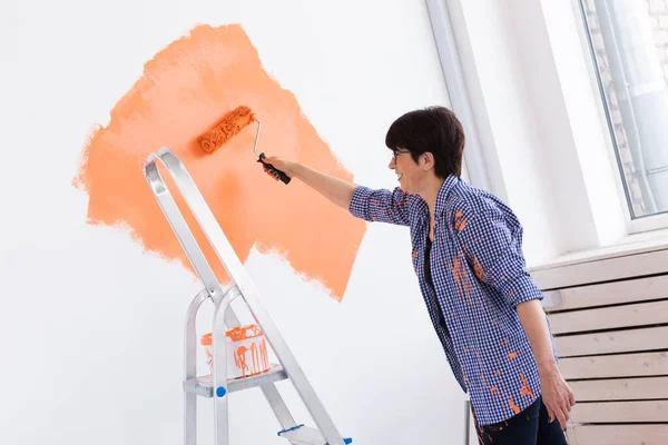 Pretty smiling middle-aged woman painting interior wall of home with paint roller. Redecoration, renovation, apartment repair and refreshment concept. — Stock Photo, Image