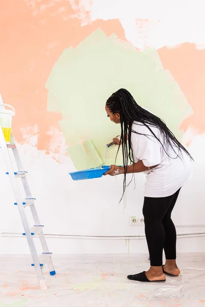 Frumoasă fată africană americană pictând peretele cu role de vopsea. Portretul unei tinere femei frumoase pictând perete în noul ei apartament. Conceptul de redecorare și renovare . — Fotografie, imagine de stoc