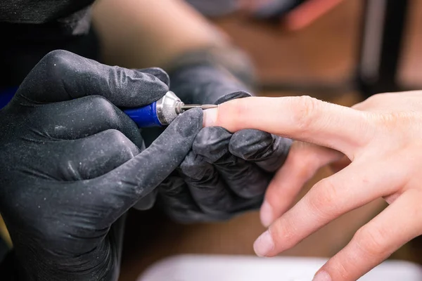 Närbild professionell kosmetolog händer arbetar med elektrisk borr på klientfingernaglar. Förfarande tillämpa artificiella naglar. Manicurist med verktyg. Manicurist i handskar tar bort nagelband — Stockfoto