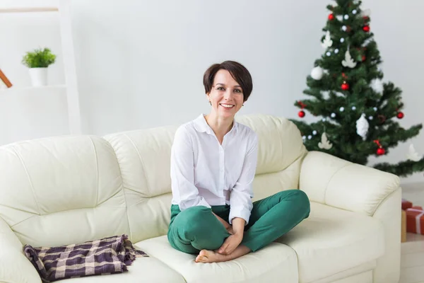 Happy woman on sofa. Christmas tree with presents under it. Decorated living room