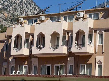 Balkonları ve güneş panelleri olan bej renkli bir bina kayalık dağ yamaçlarının önünde duruyor. Mimarlık, kentsel konutlar ve konut tasarımı açık hava manzarasıyla harmanlanmış.