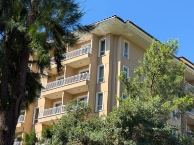 Beyaz balkonlu çok katlı bir bina kısmen uzun yeşil çam ağaçlarının arkasında gizlidir. Kentsel mimari, konut ve konut yapılarının doğal çevreyle etkileşimi