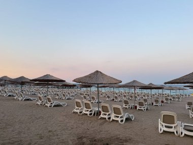 Sıralar dolusu güneş şezlongları ve saman şemsiyeleri olan kumlu bir sahil. Turizm, yaz tatili ve kıyı tatili..
