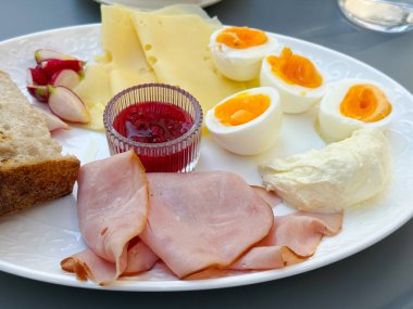 Jambonlu, peynirli, yumurtalı, ekmekli ve reçelli bir kahvaltı tabağı. Beslenme, yaşam tarzı ve yemek kültürü, sabah yemeği, gelenek ve dengeli gıda seçeneklerini göstermektedir.