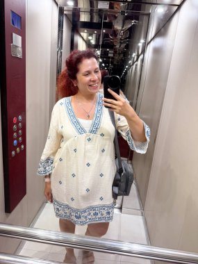 Asansörde aynalı selfie çeken gülümseyen kadın sıradan beyaz elbiseler giyiyor. Yaşam tarzı, moda ve günlük ruh hali bireyselliği, stili ve neşeli varlığı ifade eder.