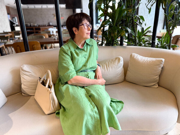 Woman in green dress sitting on sofa with handbag. Lifestyle, leisure, and modern relaxation indoors