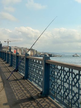 Balıkçı oltası köprü korkuluklarına yaslanıyor. Arka planda deniz ve tekneler var. Kentsel yaşam tarzı, boş zaman ve şehir ortamında geleneksel aktivite