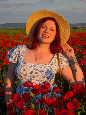 Gün batımında kırmızı gelinciklerle çevrili hasır şapkalı bir kadın gülümsüyor. Yaşam tarzı, güzellik ve kırsal kesimin doğal ahengi ile insan varlığı ve neşesi