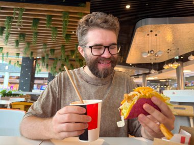 Bir adam kafe masasında taco tutarken pipetle bardaktan içer. Tüketici yaşam tarzı, fast food kültürü, gastronomi ve modern şehir ortamında günlük yemek.