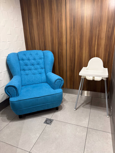 Armchair and baby highchair placed in tiled interior corner. Family care, comfort, household furnishing and public facility with parenting lifestyle and supportive design