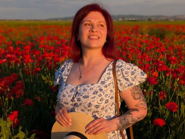 Gün batımında kırmızı gelinciklerle çevrili hasır şapkalı bir kadın gülümsüyor. Yaşam tarzı, güzellik ve kırsal kesimin doğal ahengi ile insan varlığı ve neşesi