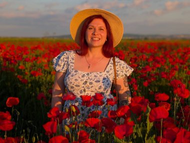 Gün batımında kırmızı gelinciklerle çevrili hasır şapkalı bir kadın gülümsüyor. Yaşam tarzı, güzellik ve kırsal kesimin doğal ahengi ile insan varlığı ve neşesi