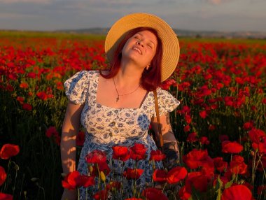 Gün batımında kırmızı gelinciklerle çevrili hasır şapkalı bir kadın gülümsüyor. Yaşam tarzı, güzellik ve kırsal kesimin doğal ahengi ile insan varlığı ve neşesi