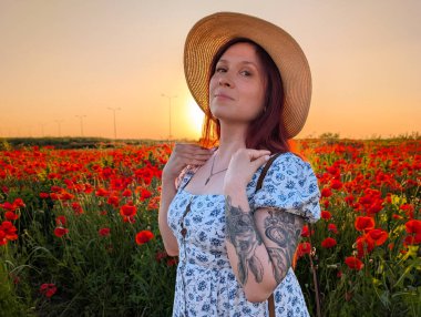 Gün batımında kırmızı gelinciklerle çevrili hasır şapkalı bir kadın gülümsüyor. Yaşam tarzı, güzellik ve kırsal kesimin doğal ahengi ile insan varlığı ve neşesi