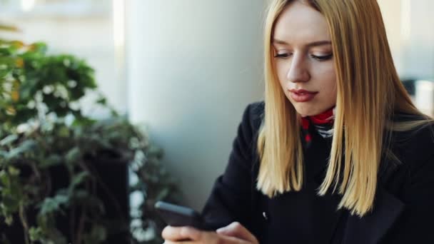 Mladá blonďatá dívka pracuje s smartphone sittig u stolu v kavárně — Stock video