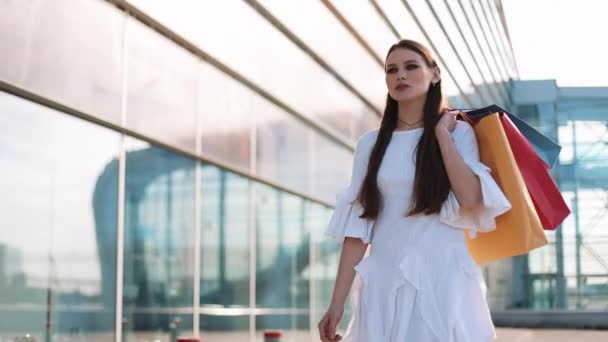 Modelo de moda bonita em poses vestido branco com sacos de compras antes de um edifício de vidro moderno. Movimento lento — Vídeo de Stock