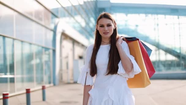 Mooie mannequin in witte jurk vormt met boodschappentassen voordat een moderne glazen gebouw — Stockvideo