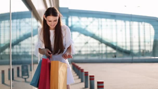 Modelo de moda bonita no vestido branco posa com sacos de compras antes de um edifício de vidro moderno — Vídeo de Stock