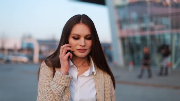 Молодая женщина с длинными волосами ходит по улице и разговаривает по телефону — стоковое видео