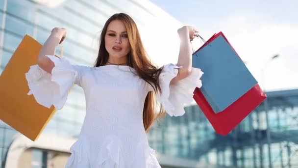 Jolie mannequin en robe blanche pose avec des sacs à provisions devant un bâtiment en verre moderne — Video