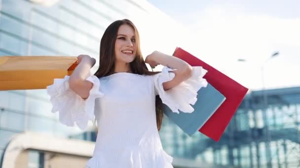 Прелестная модель в белом платье позирует с сумками перед современным стеклянным зданием — стоковое видео