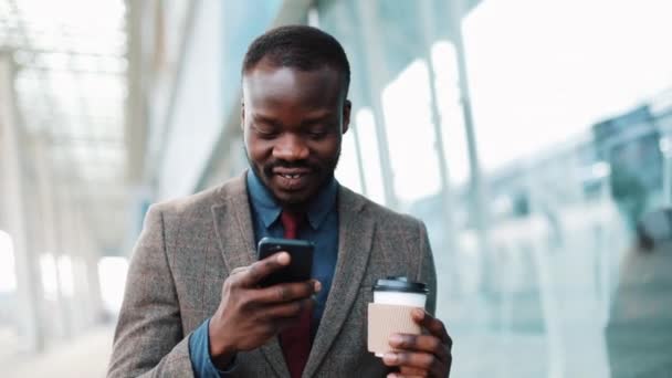 Hombre afroamericano escribe algo en su teléfono inteligente caminando fuera. El hombre de negocios camina por la terminal del aeropuerto, usa su teléfono, bebe café, imprime el mensaje, sonríe. Alegre humor, ser feliz — Vídeo de stock