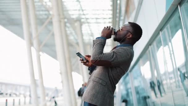 Heureux homme d'affaires afro-américain écoutant de la musique dans les écouteurs sur smartphone, marchant en dehors du bureau et dansant drôle au rythme. Buvez du café, dansez bizarrement. Gros plan — Video