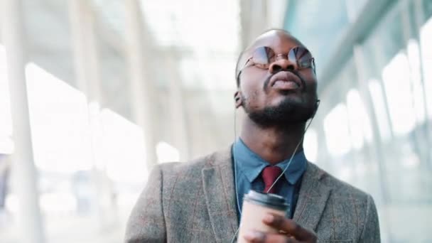 Feliz hombre de negocios afroamericano escuchando música en auriculares en el teléfono inteligente, caminando fuera de la oficina y bailando divertido al ritmo. Beber café, divertidos movimientos de baile. Primer plano, cámara lenta — Vídeo de stock