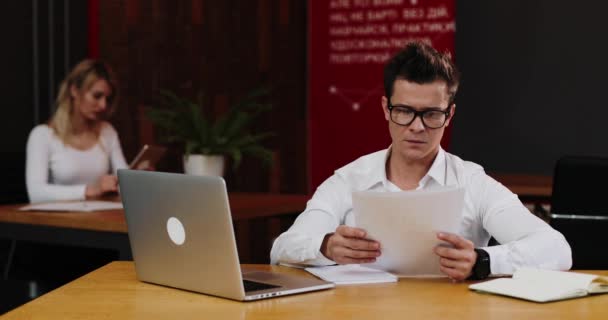 Joven hombre de negocios en gafas con ordenador portátil y papel de trabajo en la oficina. Negocios, personas, papeleo y concepto de tecnología. 4k, cámara lenta — Vídeos de Stock