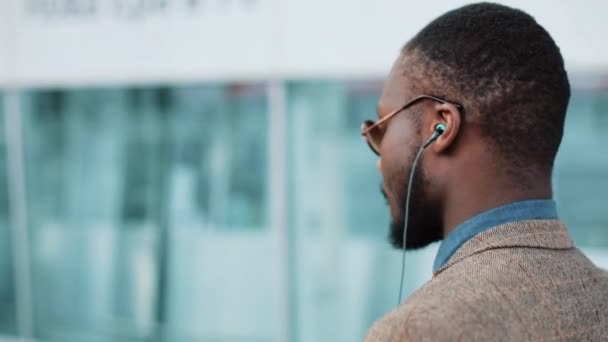 Empresário afro-americano ouve música e dança enquanto caminha perto do prédio de escritórios com um smartphone. Conceito: trabalhador de escritório, descanso, publicidade — Vídeo de Stock