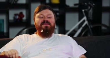 Sakallı şişman adam gülümseyen odasında Tv izliyor ve bira ile masada mısır yiyor. Şişman adam bira içiyor. Konsept: Yetersiz beslenme, sedanter yaşam tarzı, dinlenme kanepeye, obezite