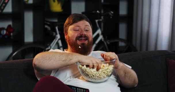 Dikke man met baard glimlachend horloges Tv op de kamer en eet pop-maïs aan tafel met bier. Dikke man drinkt bier. Concept: Ondervoeding, sedentaire levensstijl, rusten op de Bank, obesitas — Stockvideo