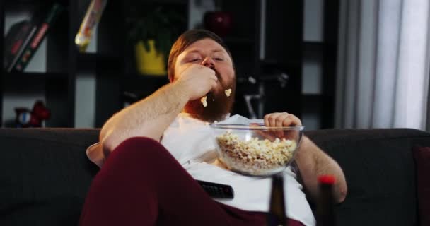 Homem gordo sorridente com barba assiste TV no quarto e come pipoca na mesa com cerveja. O gordo bebe cerveja. Conceito: Desnutrição, sedentarismo, descanso no sofá, obesidade — Vídeo de Stock